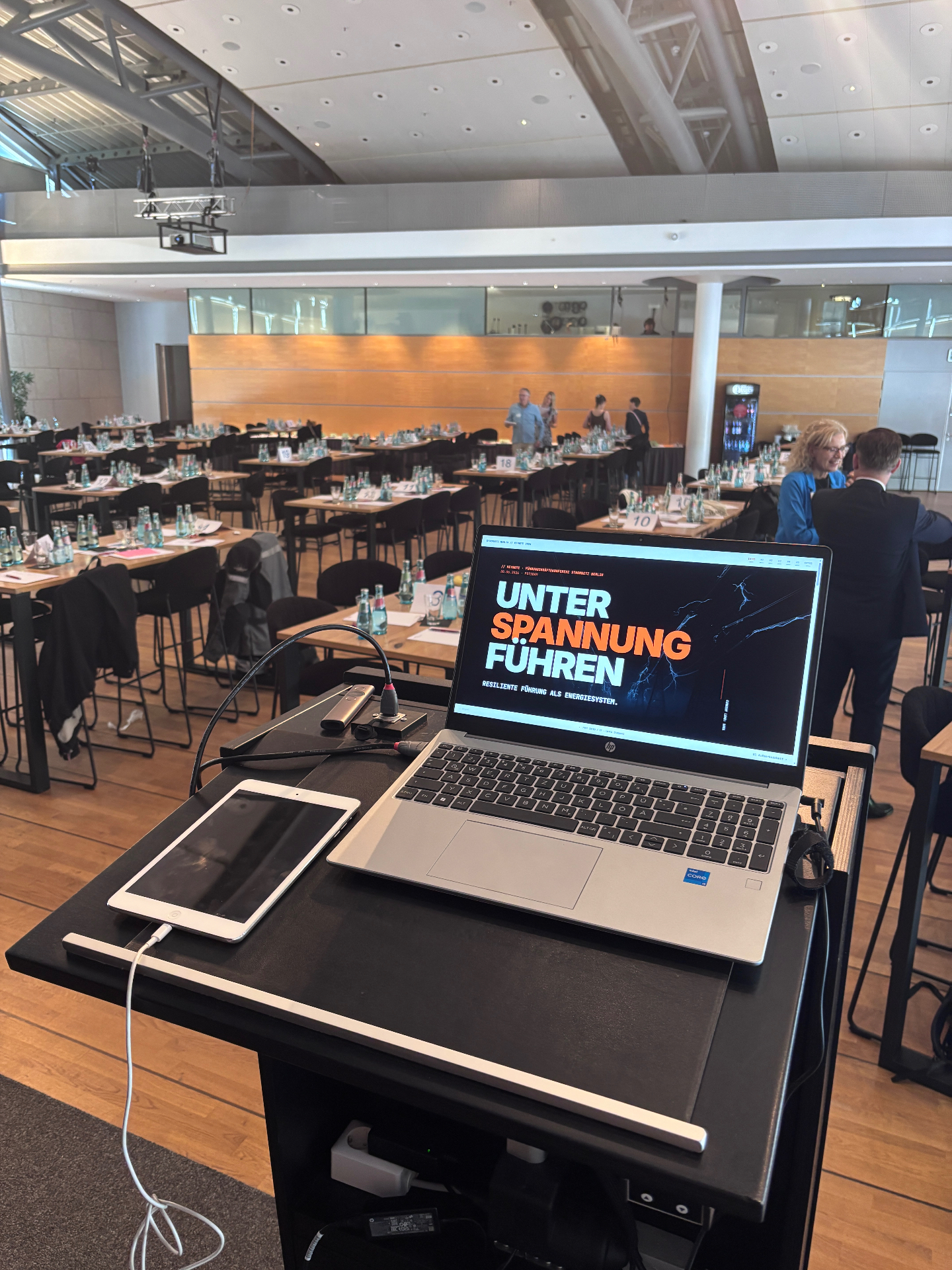 Rednerpult mit Laptop, auf dem Bildschirm die Keynote-Titelfolie „Unter Spannung führen“, im Hintergrund der Konferenzsaal in Potsdam